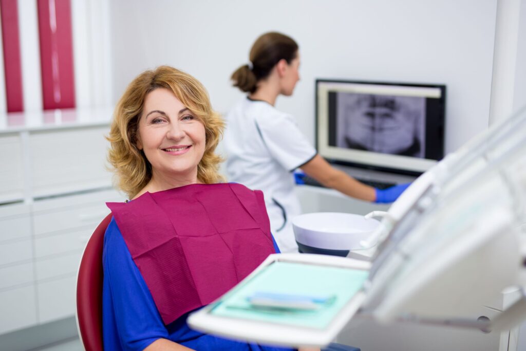 Woman in blue shirt in dental chair smiling with dentist in background with X-rays